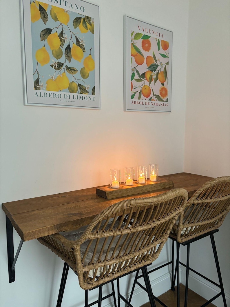 May include: A cozy dining area featuring a wooden table with two woven rattan chairs. Above, two framed botanical prints depict lemons and oranges. A wooden tray holds five lit candles, creating a warm ambiance.