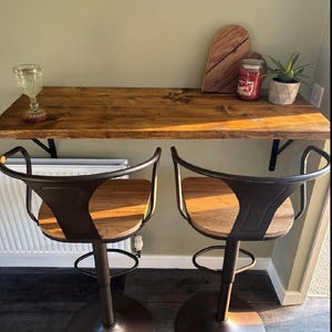 May include: Two brown metal bar stools with wooden seats are positioned in front of a wooden bar. The bar has a glass, a wooden cutting board, a candle, and a small potted plant. The stools have a circular base and a curved back.