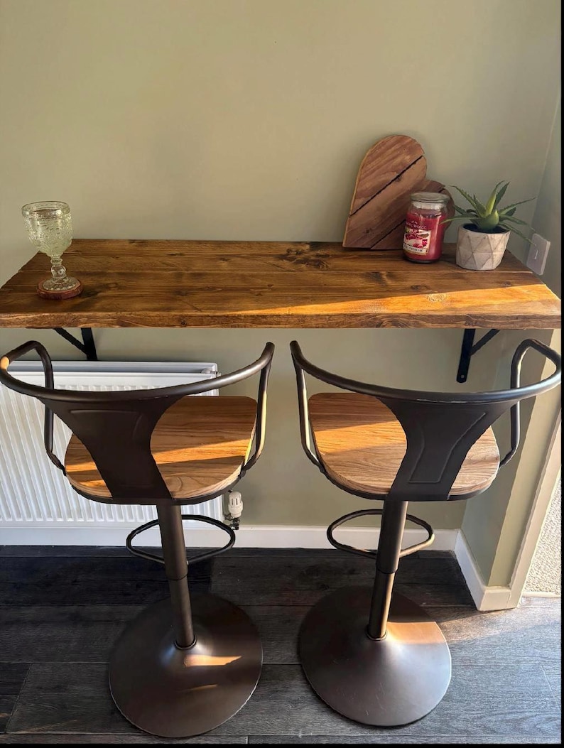 May include: Two brown metal bar stools with wooden seats are positioned in front of a wooden bar. The bar has a glass, a wooden cutting board, a candle, and a small potted plant. The stools have a circular base and a curved back.