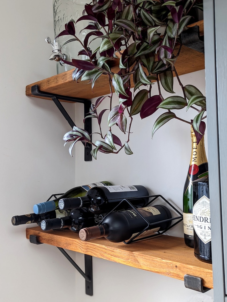 May include: A wooden shelf with black metal brackets holds a wine rack with several bottles of wine and a bottle of champagne. A green plant with purple leaves hangs down from the shelf.