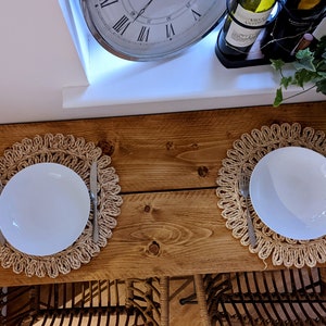 May include: Two white plates, each set on a woven placemat, with a fork and knife. The table is made of wood. A clock and wine bottles are visible in the background. The scene suggests a dining setting.