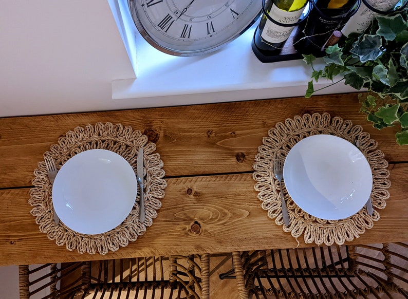 May include: Two white plates, each set on a woven placemat, with a fork and knife. The table is made of wood. A clock and wine bottles are visible in the background. The scene suggests a dining setting.