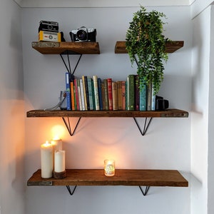 May include: Three wooden shelves with black metal brackets, decorated with books, candles, and vintage cameras. The shelves are mounted on a white wall.