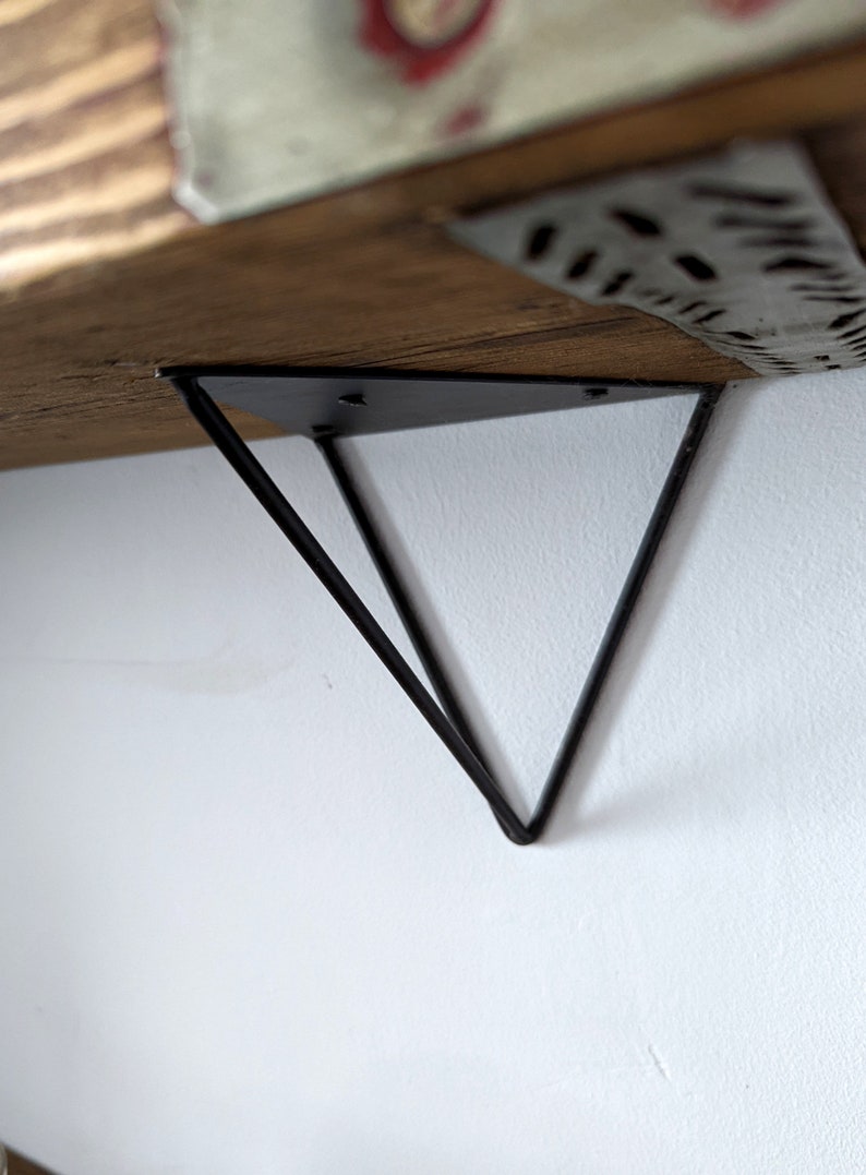 May include: Black metal shelf bracket with a triangular design. The bracket is attached to a wooden shelf.