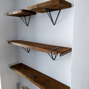 May include: Four wooden shelves with black metal triangular brackets mounted on a white wall. The shelves are made of reclaimed wood and have a rustic look.