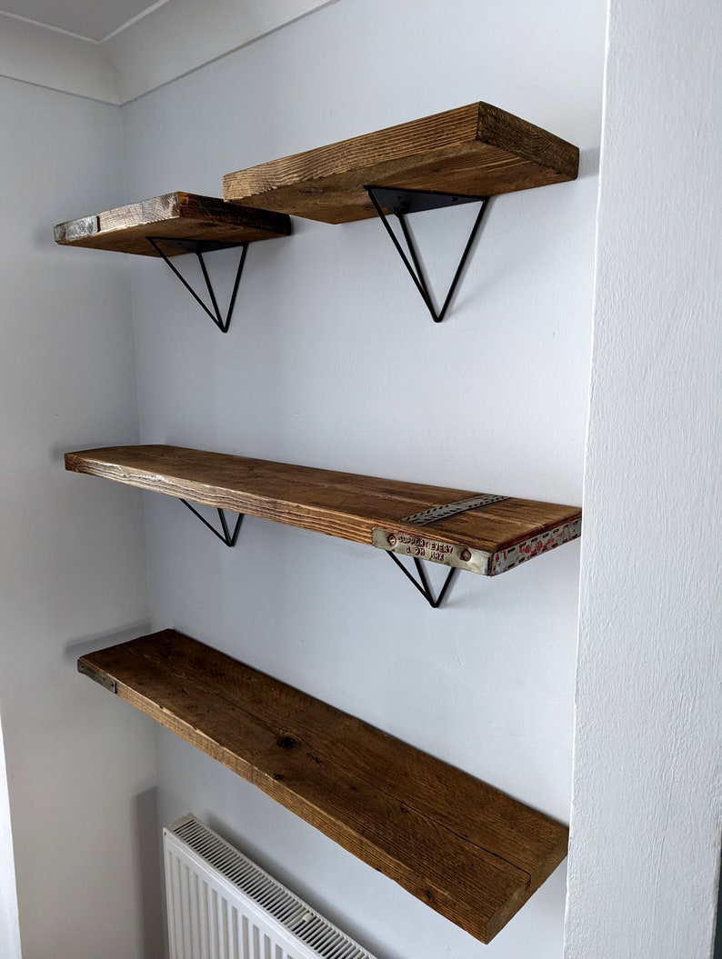 May include: Four wooden shelves with black metal triangular brackets mounted on a white wall. The shelves are made of reclaimed wood and have a rustic look.