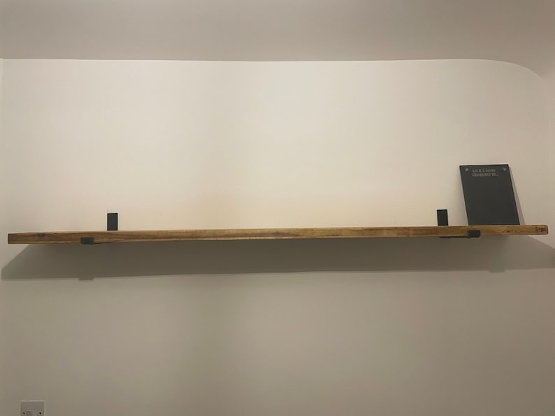 May include: A wooden shelf with black metal brackets. The shelf is mounted on a white wall. A small black chalkboard with the text "Just Love Remember It" is sitting on the shelf.