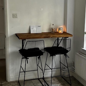 May include: A rustic wooden bar table with two black metal bar stools. The stools have black cushions and a unique backrest design. A small candle holder and a bottle are on the table. The setup is against a white wall.