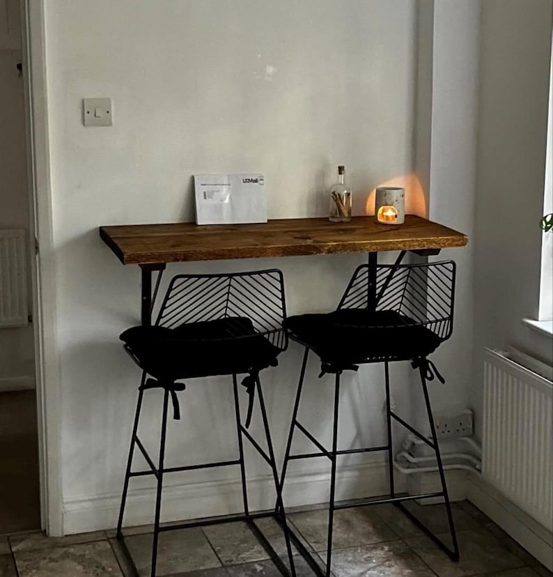 May include: A rustic wooden bar table with two black metal bar stools. The stools have black cushions and a unique backrest design. A small candle holder and a bottle are on the table. The setup is against a white wall.