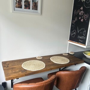 May include: A rustic, wall-mounted wooden table with two brown leather chairs. The table has two woven placemats and two matching coasters. A framed picture of three dogs hangs on the wall above the table. A chalkboard with handwritten notes is also visible.