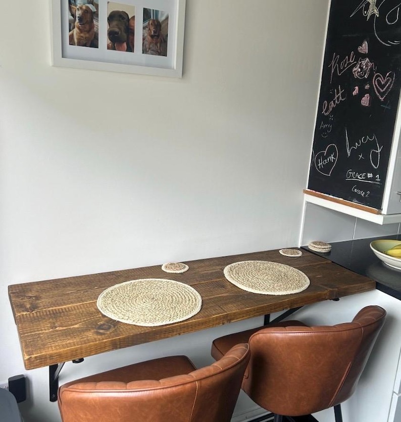 May include: A rustic, wall-mounted wooden table with two brown leather chairs. The table has two woven placemats and two matching coasters. A framed picture of three dogs hangs on the wall above the table. A chalkboard with handwritten notes is also visible.