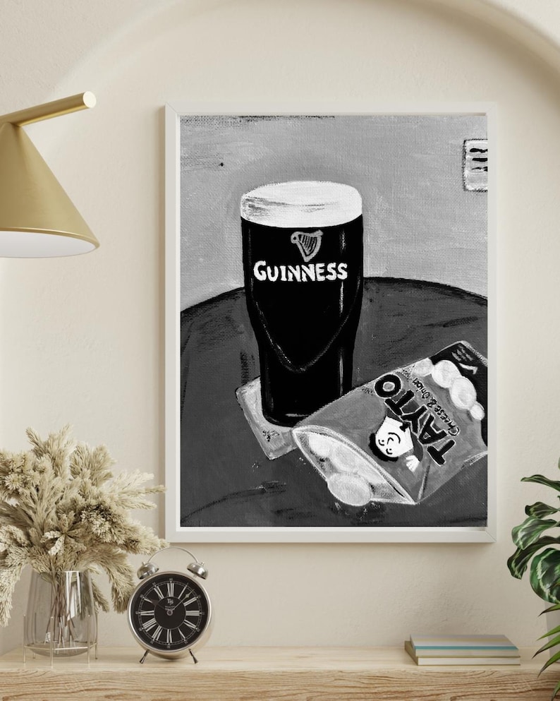 Peut inclure: Impression en noir et blanc d'une pinte de Guinness avec le logo Guinness et un paquet de chips Tayto au fromage et &agrave; l'oignon. L'&oelig;uvre est encadr&eacute;e en blanc.