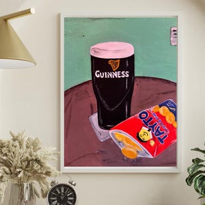 Peut inclure: Impression d'art encadr&eacute;e repr&eacute;sentant un verre de Guinness avec une mousse rose et le logo Guinness, &agrave; c&ocirc;t&eacute; d'un paquet rouge de chips Tayto au fromage et &agrave; l'oignon. Fond vert et marron.