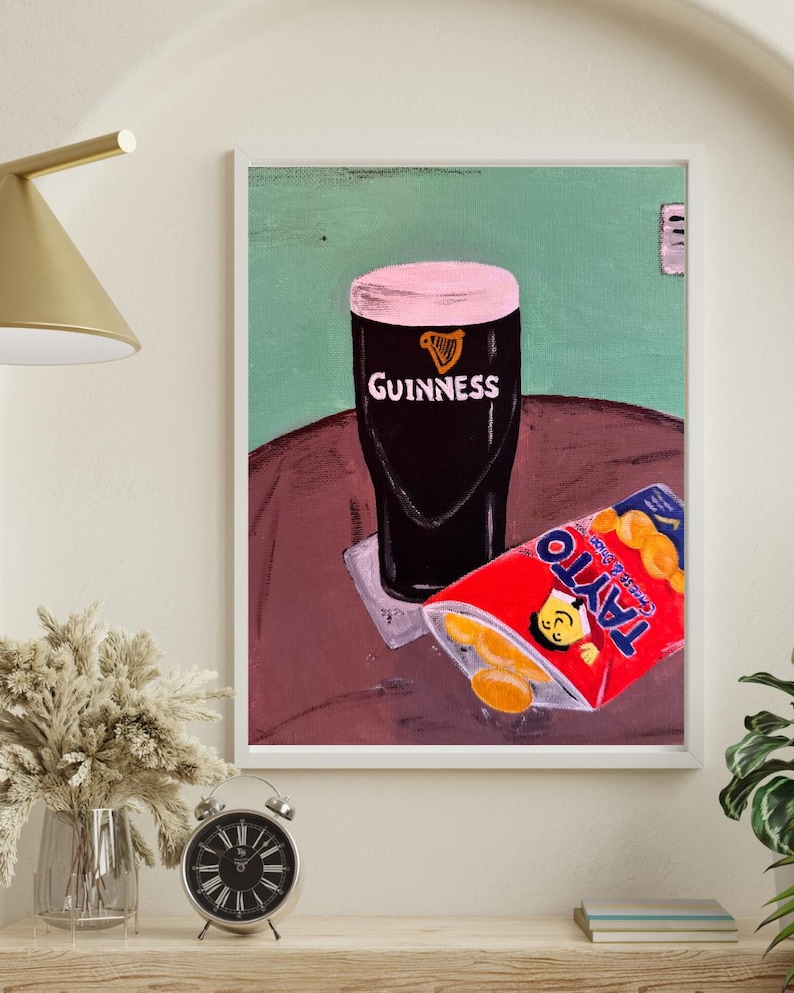 Peut inclure: Impression d'art encadr&eacute;e repr&eacute;sentant un verre de Guinness avec une mousse rose et le logo Guinness, &agrave; c&ocirc;t&eacute; d'un paquet rouge de chips Tayto au fromage et &agrave; l'oignon. Fond vert et marron.