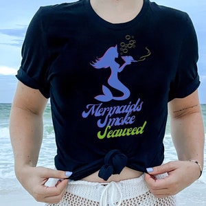 May include: A black t-shirt with a purple mermaid smoking a pipe and the text "Mermaids Smoke Seaweed" in a colorful font.