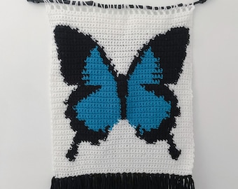 Peut inclure: Une tapisserie murale au crochet représentant un papillon bleu avec des ailes noires. La tapisserie a un fond blanc et une frange noire.