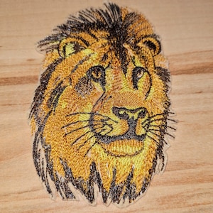 Puede incluir: Parche bordado de cabeza de león con melena negra y pelaje amarillo. Los ojos del león son marrones y la nariz es negra.