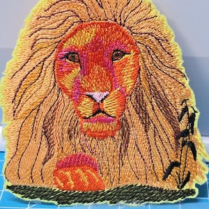 Puede incluir: Parche bordado de un león con una melena naranja y amarilla detallada, una nariz rosa y un contorno negro. El león mira directamente al espectador. El parche está sobre un fondo amarillo.