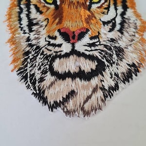 Può includere: Primo piano di una toppa ricamata con il viso di una tigre. La tigre ha strisce arancioni e nere, un muso bianco e occhi gialli.