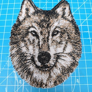 Könnte beinhalten: Gestickter Wolfskopf-Patch mit braunem, schwarzem und weißem Fell. Der Patch befindet sich auf einer blauen Schneidematte.
