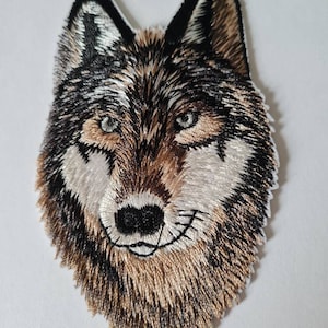 Peut inclure: Patch thermocollant brodé représentant la tête d'un loup. Le loup a une fourrure brune, noire et blanche. Les yeux du loup sont bleus et il a un nez noir.