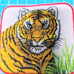 Peut inclure: Patch brodé représentant un gros plan sur le visage d'un tigre avec des yeux verts, des rayures orange et noires, et un fond blanc. Le tigre est entouré d'herbe verte.