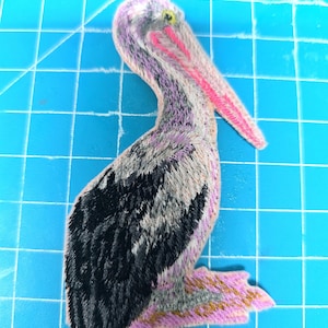 Könnte beinhalten: Gestickter Aufnäher eines Pelikans in Grau-, Schwarz-, Rosa- und Lilatönen. Der Pelikan hat einen langen rosa Schnabel und ein detailliertes Federdesign. Der Aufnäher befindet sich auf einem blauen Gitterhintergrund.