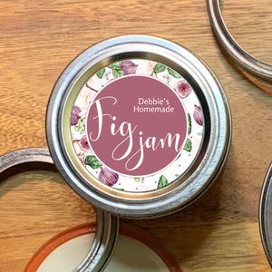 Peut inclure: Un couvercle de bocal en argent avec une étiquette blanche sur laquelle est inscrit "Debbie's Homemade Fig Jam" en écriture rose. L'étiquette est ornée d'un motif d'aquarelle de figues et de feuilles.