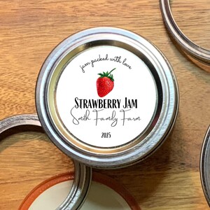 May include: A white round jar lid with a label that says "jam packed with love" and "STRAWBERRY JAM Smith Family Farm 2023". A red strawberry with green leaves is pictured on the label.