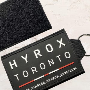 May include: Black rectangular patch with the text "HYROX TORONTO" in white, and a red and white line. The patch is attached to a black strap with a silver keyring. A black velcro patch is in the background.
