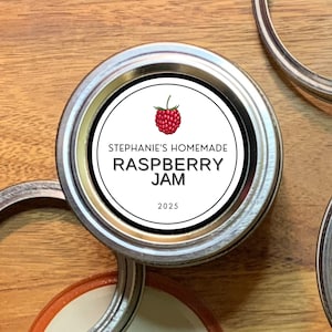 May include: A white round jar label with black text and a red raspberry illustration. The label reads "Stephanie's Homemade Raspberry Jam 2025".