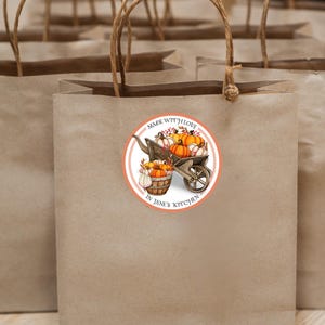 May include: A round sticker with a peach background and white border. The sticker features a hand-drawn illustration of a wheelbarrow filled with pumpkins and the text "Made with Love in Jane's Kitchen".