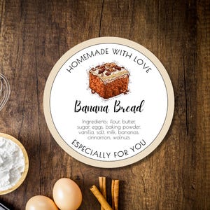 May include: A round, beige label with the text "HOMEMADE WITH LOVE" and "ESPECIALLY FOR YOU" in a brown font. The label also features a drawing of a slice of banana bread with walnuts on top and the text "Banana Bread" in a brown font. The text "Ingredients: flour, butter, sugar, eggs, baking powder, vanilla, salt, milk, bananas, cinnamon, walnuts" is also included in a smaller brown font.
