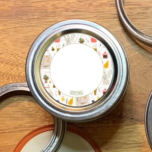 Könnte beinhalten: Ein weißer runder Aufkleber mit einem Blumenmuster und dem aufgedruckten Wort "HOMEMADE". Der Aufkleber ist auf einem silbernen Metalldeckel befestigt.