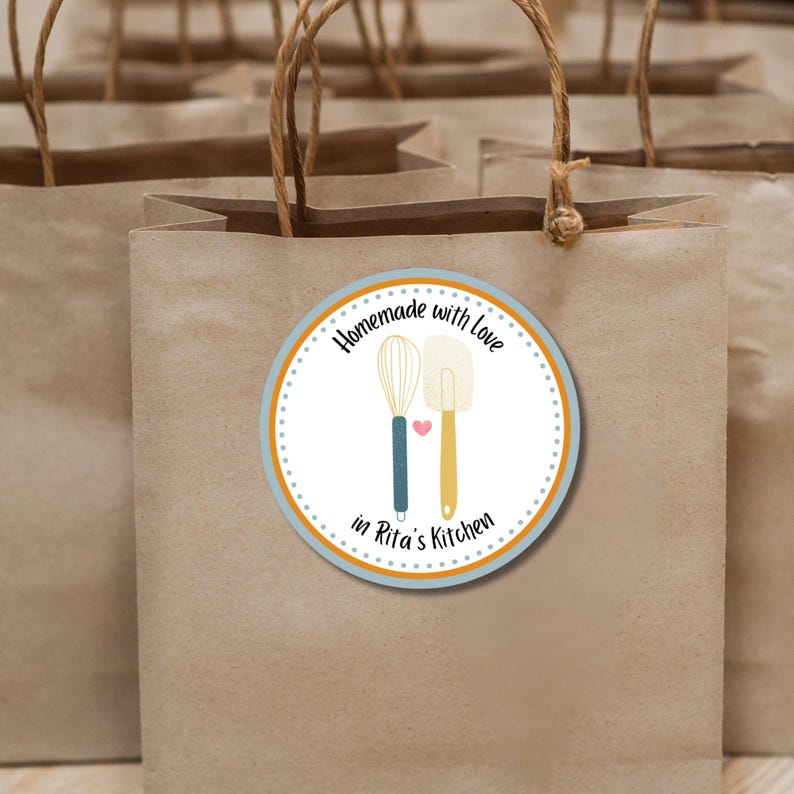 May include: A brown paper bag with a round sticker that says "Homemade with love in Rita's Kitchen". The sticker has a blue and orange border and features a whisk and a spatula with a pink heart between them.
