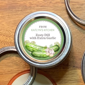 Peut inclure: Une étiquette ronde verte et blanche pour un bocal avec le texte "From Kaitlyn's Kitchen Zesty Dill with Extra Garlic". L'étiquette présente une illustration aquarelle de concombres et d'ail.