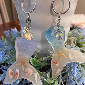 Pode incluir: Dois chaveiros com formato de cauda de sereia em resina azul e branca iridescente, com conchas, estrelas do mar e uma borboleta. Cada chaveiro tem um anel prateado e um charme de cristal.