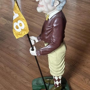 May include: A detailed figurine of a golfer holding a flag with the number 18. The figure wears a brown jacket, yellow and brown plaid cap, and khaki pants. The base is green, and the flag is yellow.