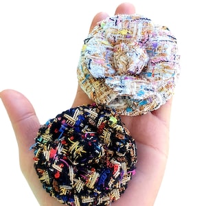 Könnte beinhalten: Zwei Stoffblumenbroschen, eine weiße mit goldenen und rosafarbenen Akzenten, die andere schwarze mit goldenen, roten und blauen Akzenten.