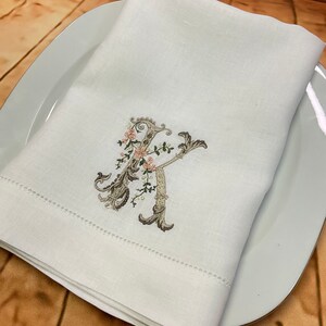 May include: White linen napkin with a delicate embroidered floral design featuring a capital letter "K" in a script font. The embroidery is in shades of brown and pink.