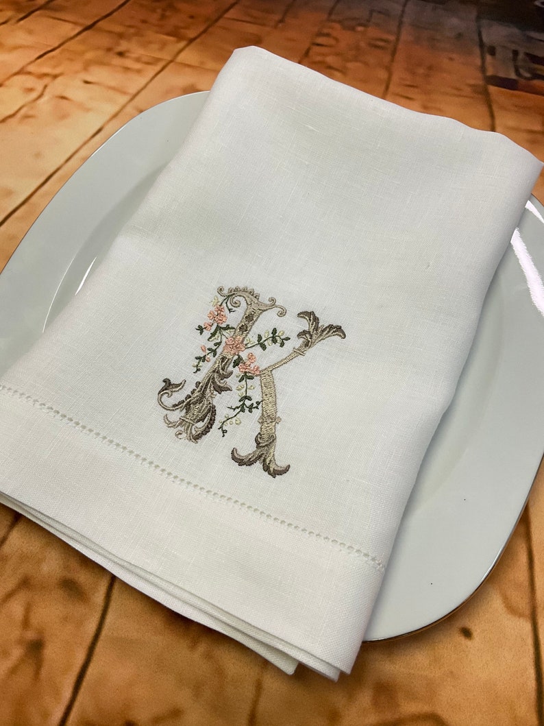 May include: White linen napkin with a delicate embroidered floral design featuring a capital letter "K" in a script font. The embroidery is in shades of brown and pink.