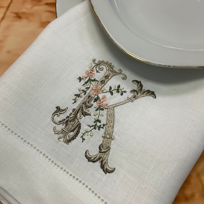 May include: A white linen napkin with a delicate embroidered floral design featuring a large letter 'R' in a brown and beige colour scheme.