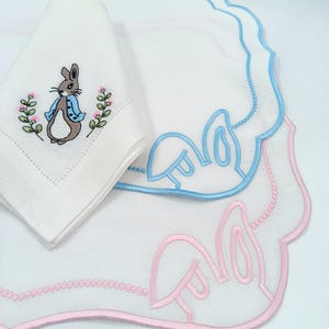 Peut inclure: Deux sets de table en lin blanc avec des motifs de lapin brodés. Le set de table du haut présente un lapin bleu portant une veste bleue et des fleurs roses. Le set de table du bas présente un lapin rose avec des oreilles roses.