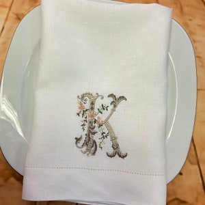 May include: White linen napkin with a monogrammed letter K embroidered in brown and green with pink floral accents.
