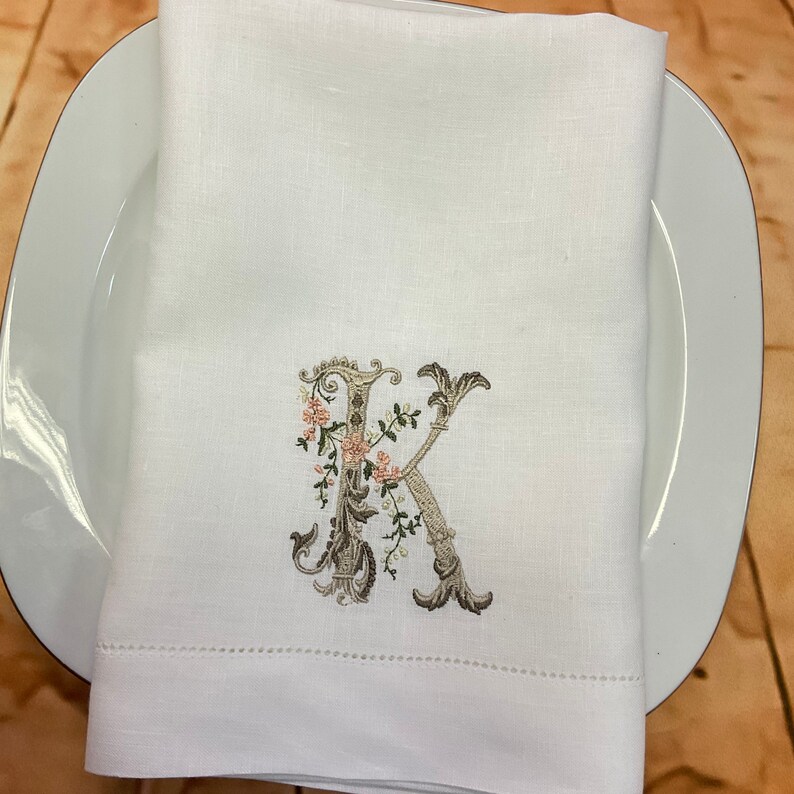 May include: White linen napkin with a monogrammed letter K embroidered in brown and green with pink floral accents.