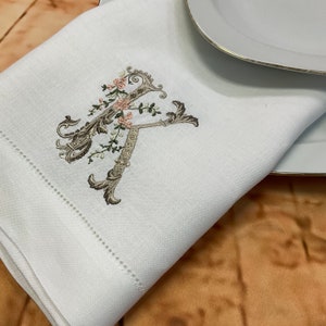 May include: White linen napkin with a delicate embroidered floral design featuring a large letter 'K' in a brown and green colour scheme.