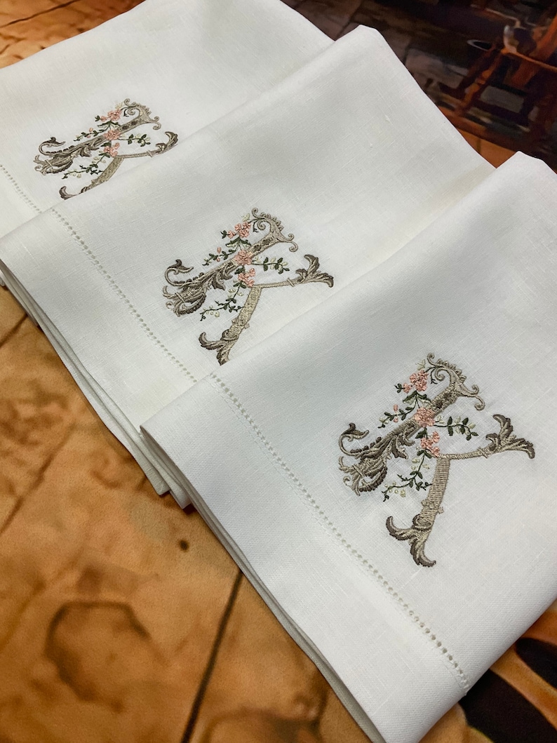 May include: Three white linen tea towels with embroidered floral designs and the letter 'A' in a script font.