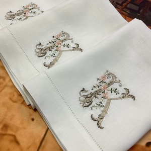 May include: Three white linen guest towels with embroidered floral monograms in shades of brown, green, and pink.