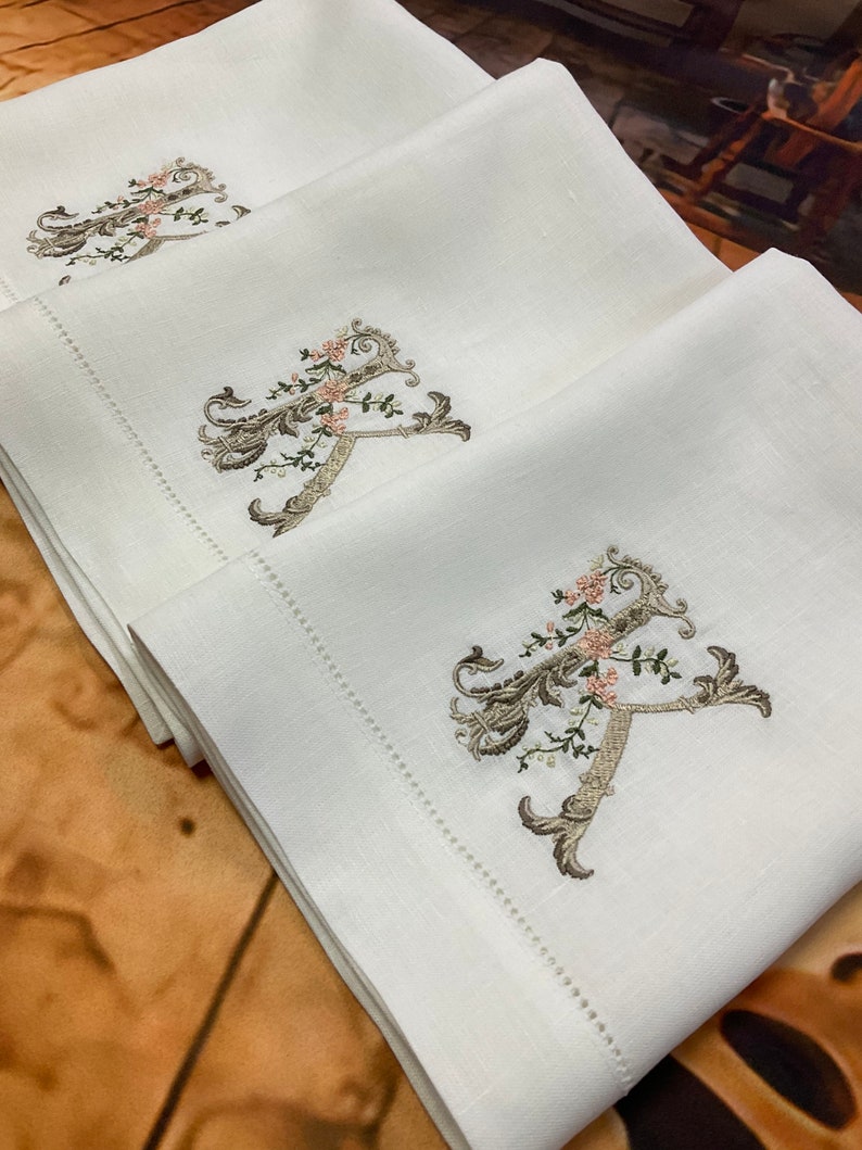 May include: Three white linen guest towels with embroidered floral monograms in shades of brown, green, and pink.