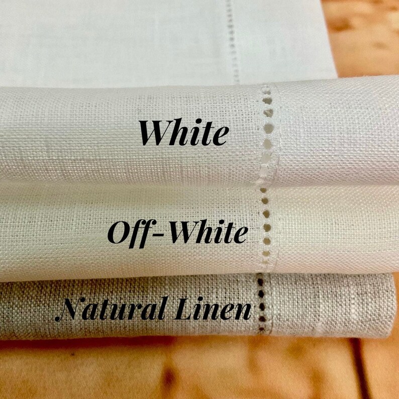 May include: Three linen napkins in different shades of white: white, off-white, and natural linen. The napkins are folded and stacked on top of each other.